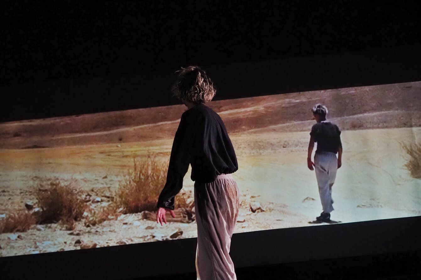 A person stands in front of a screen depicting a person walking through sand