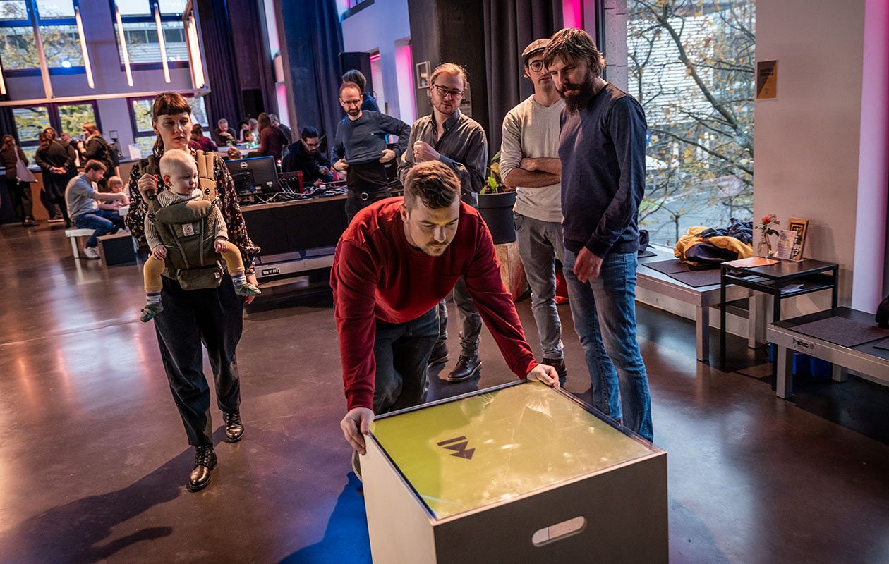 A person bends over a knee-high cube. Other people are watching him.