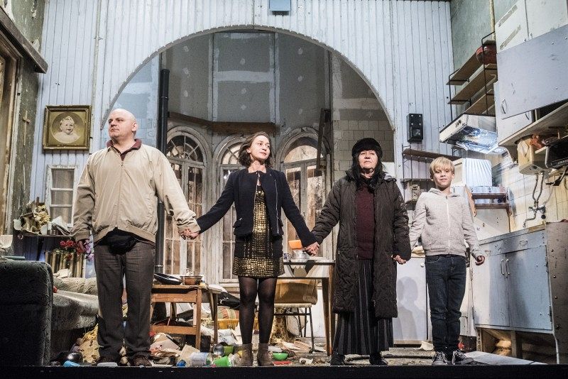 Four people hold hands and stand in front of a scenery of run down furniture.
