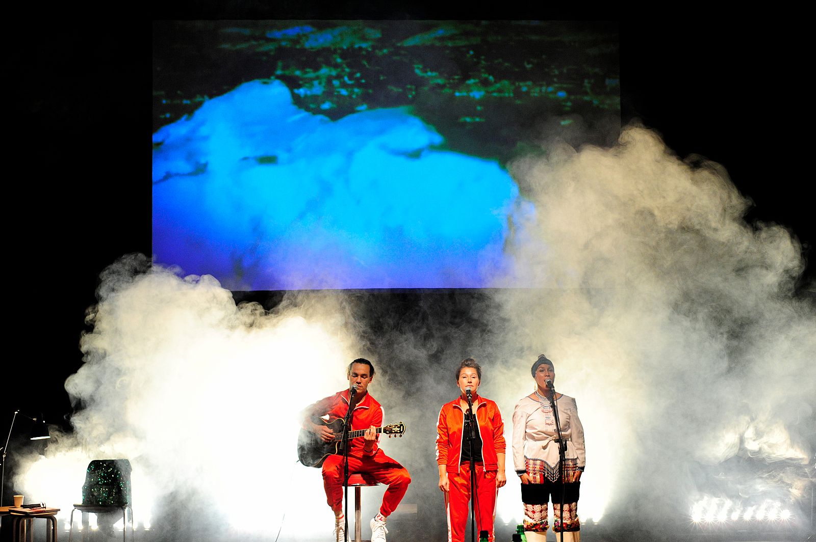 Three people with musical instruments and microphones stand on a stage with large clouds of smoke.