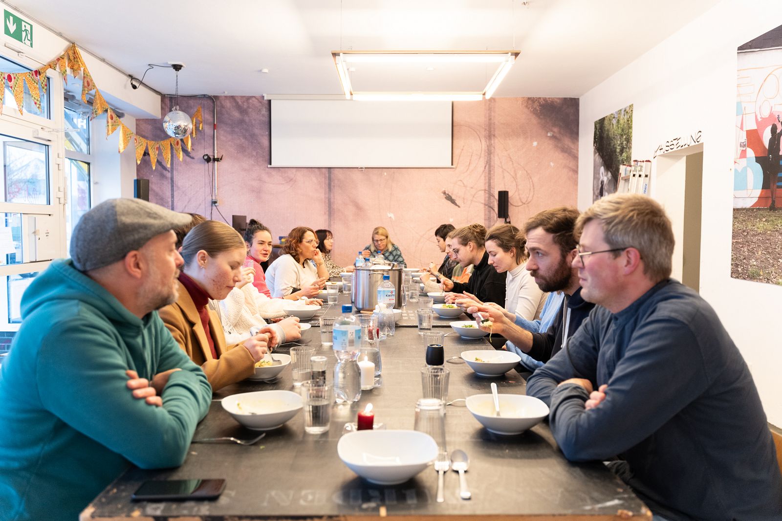 Menschen sitzen an einer langen Tafel und essen gemeinsam Suppe in einem hellen, gemütlich dekorierten Raum.