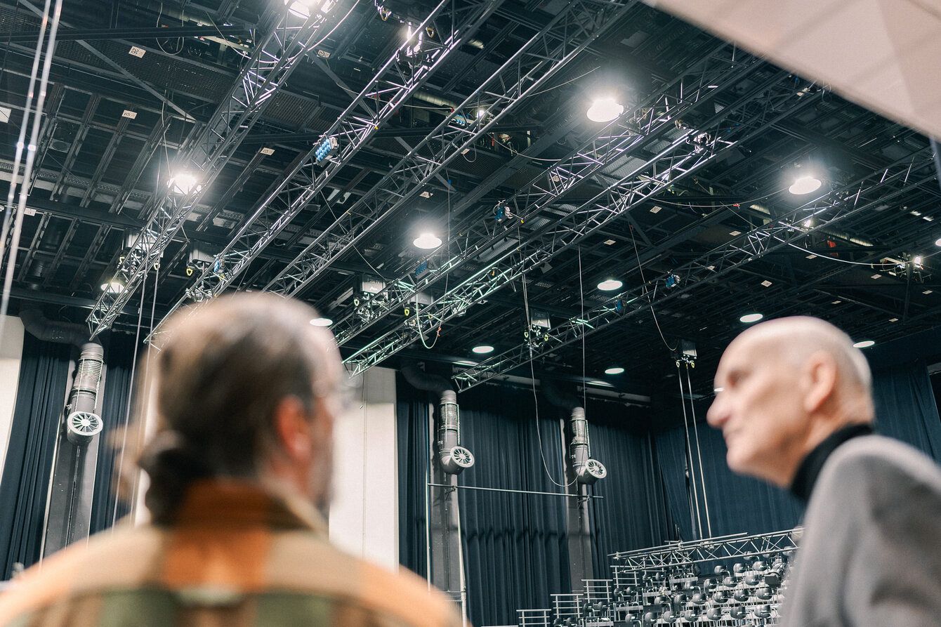 Two heads blurred in the foreground, a theatre hall with stage equipment in the background.