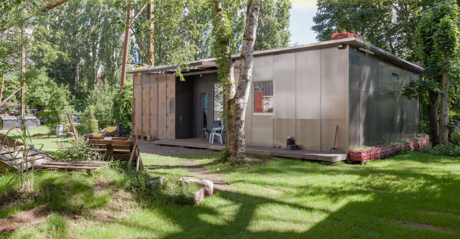 Ein einfaches Gebäude mit Blech- und Holzfassade steht in einem grünen Garten mit Bäumen, Rasenfläche und Paletten.