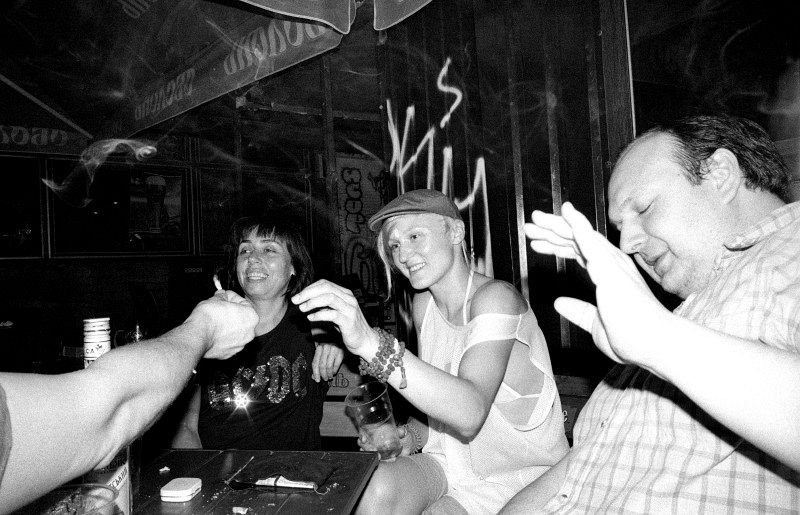 Drei Menschen, die an einem Tisch sitzen, auf dem Alkohol steht. Einer Person wird eine Zigarette hingehalten.