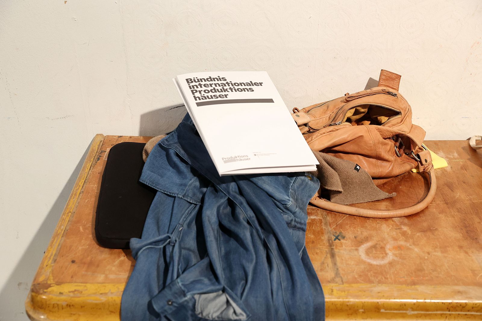 Auf einem Tisch liegt eine Reisetasche, eine Jacke und darauf eine Mappe mit dem Logo vom Bündnis internationaler Produktionshäuser