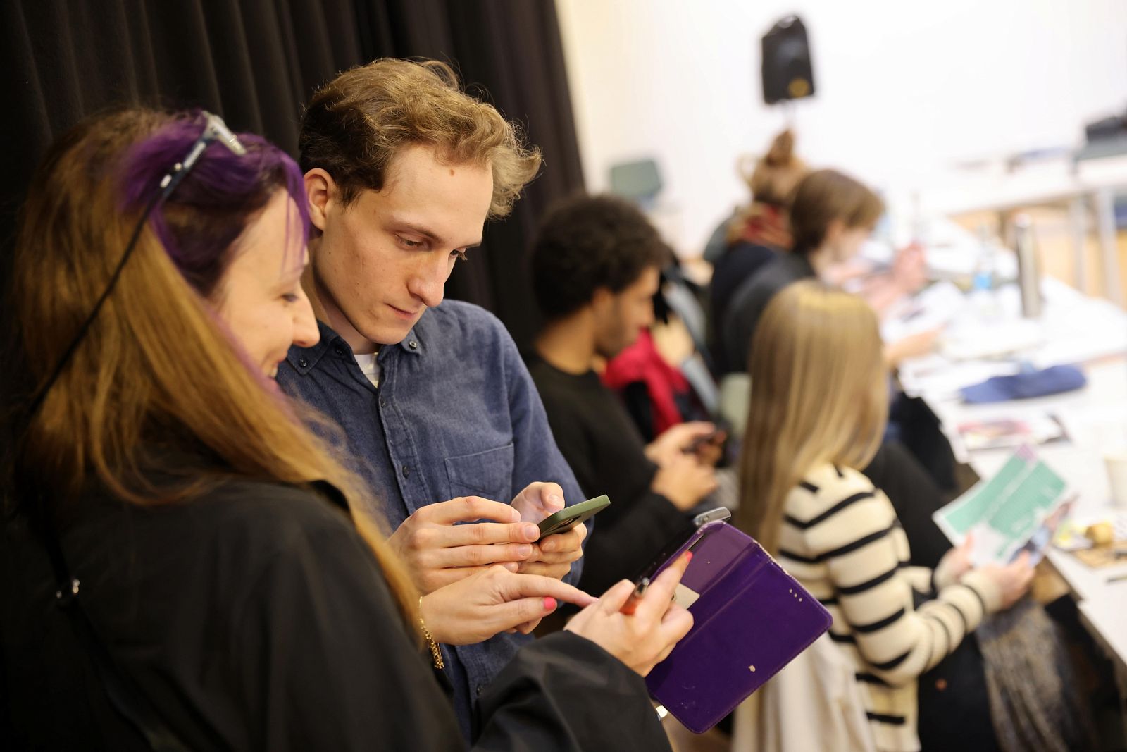 Two people in the foreground are looking intently at their smartphones; in the background, other people are sitting at a table reading.