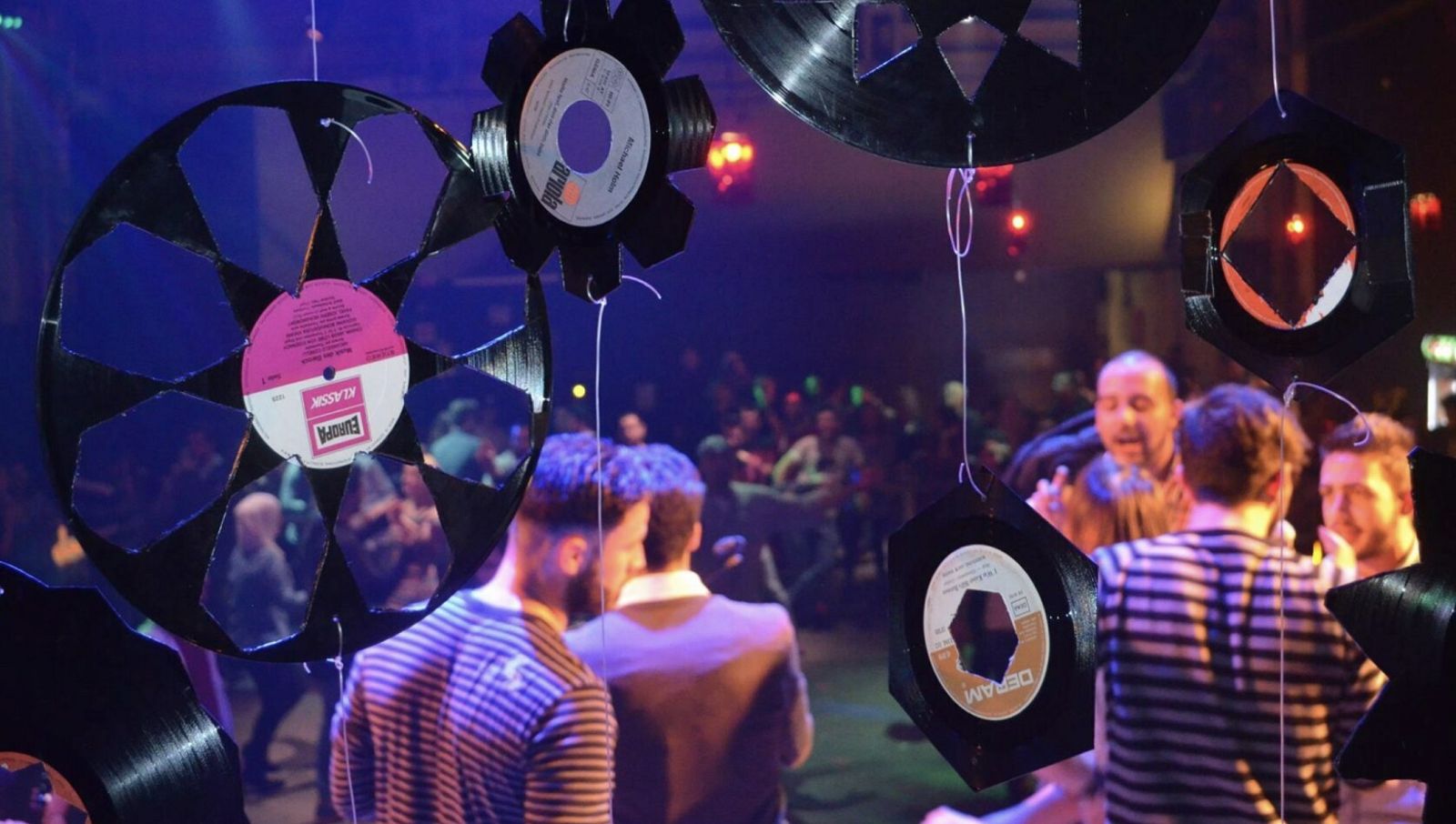 People dance in a colorfully lit room and decoratively cut-up records hang from the ceiling in the foreground.