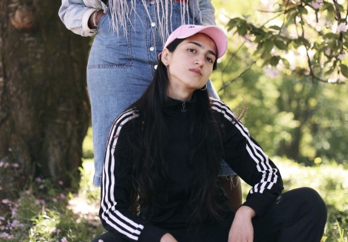 Eine junge Frau in schwarzem Trainingsanzug und rosa Mütze hockt vor einer Person in Jeanskleid. Beide sind von Bäumen und Natur umgeben.