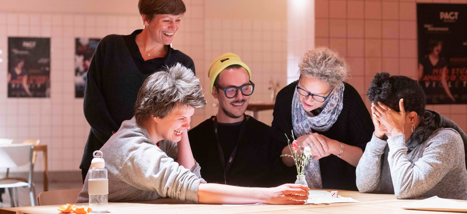 5 Personen sitzen und stehen an einem Tisch. Sie notieren nicht sichtbare Dinge auf einem Papier. Sie lachen.