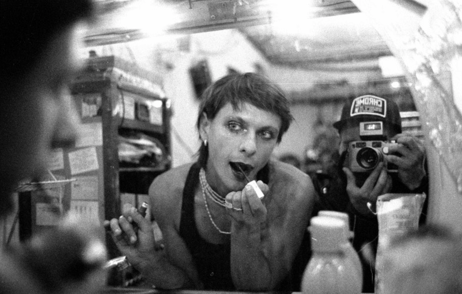 A person in front of a mirror applying lipstick and being photographed.