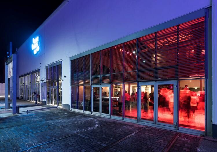 The building has large windows. Inside, people dance in the red light and a blue sign shines outside.