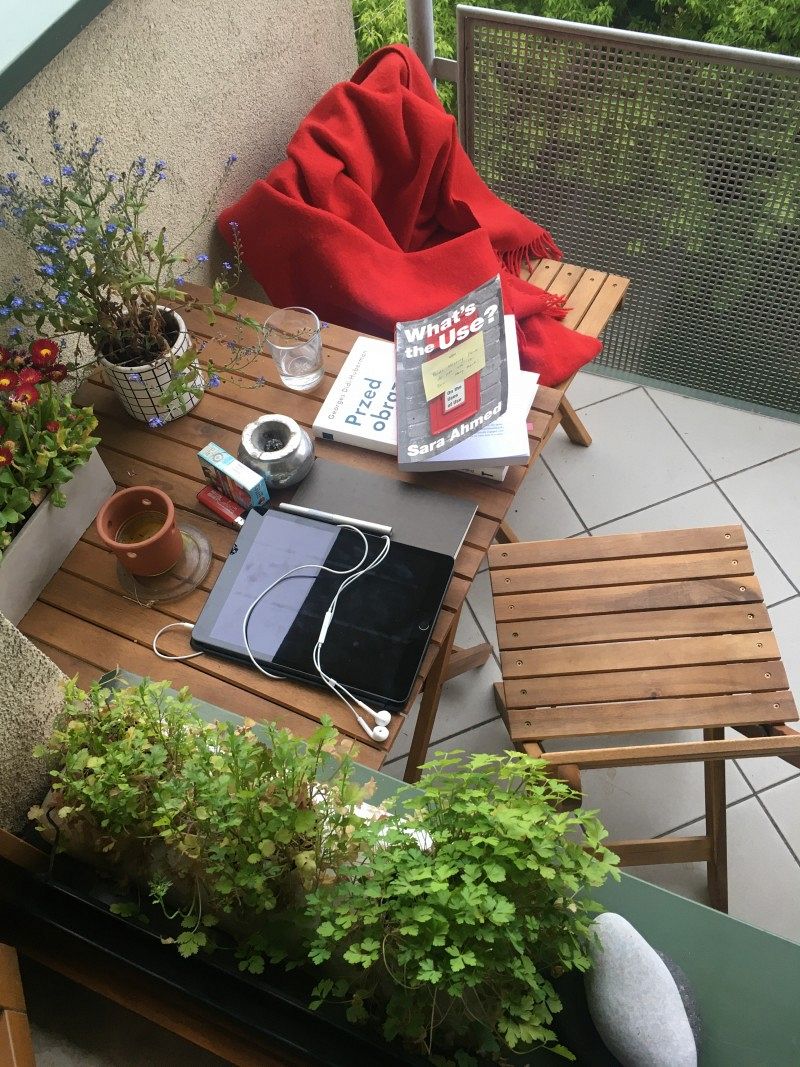 A cozy balcony seating area with a wooden table on which there are books, a gas water and a tablet, among other things.