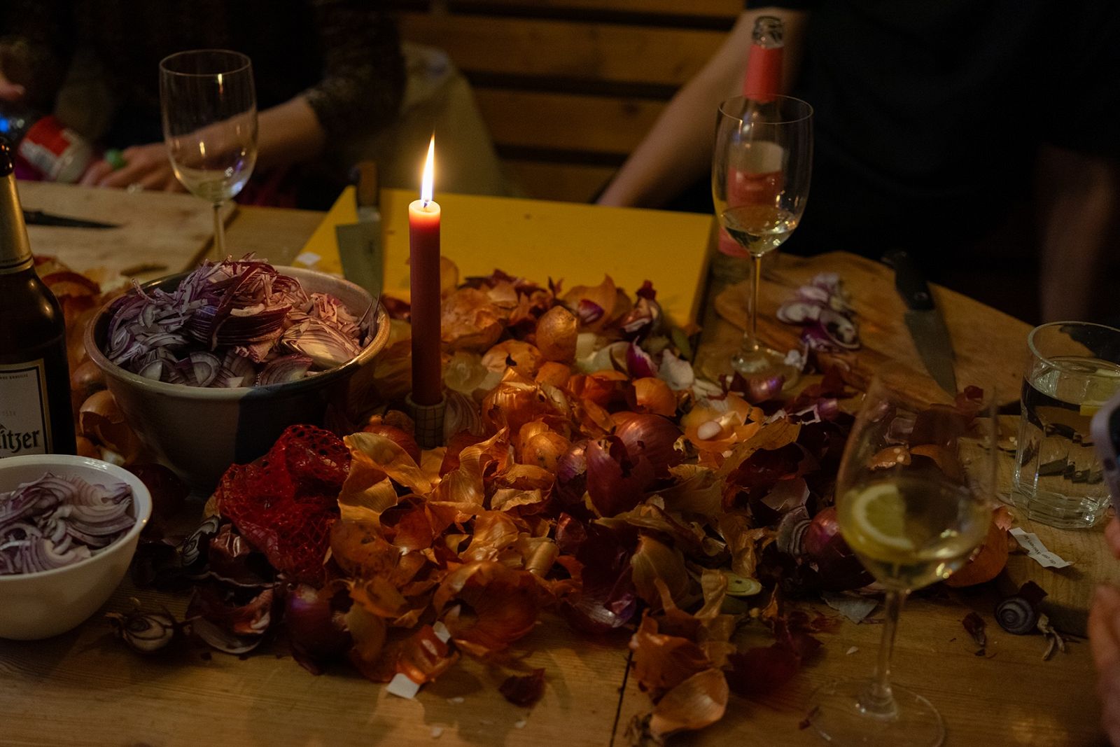 Ein Holztisch ist bedeckt mit Zwiebelschalen, geschnittenen roten Zwiebeln, Weingläsern, einer brennenden Kerze und Küchenutensilien – eine gemütliche Vorbereitungsrunde.