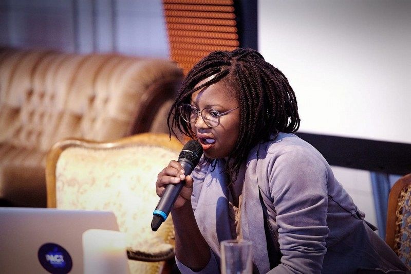 A woman with a microphone in her hand, sitting on a chair with a computer in front of her.