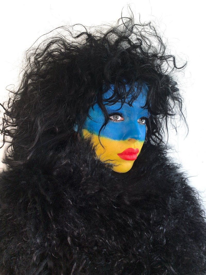 Eine Frau mit dunklen Locken und dunkler Jacke, deren Gesicht mit einer Ukraine-Flagge bemalt ist.